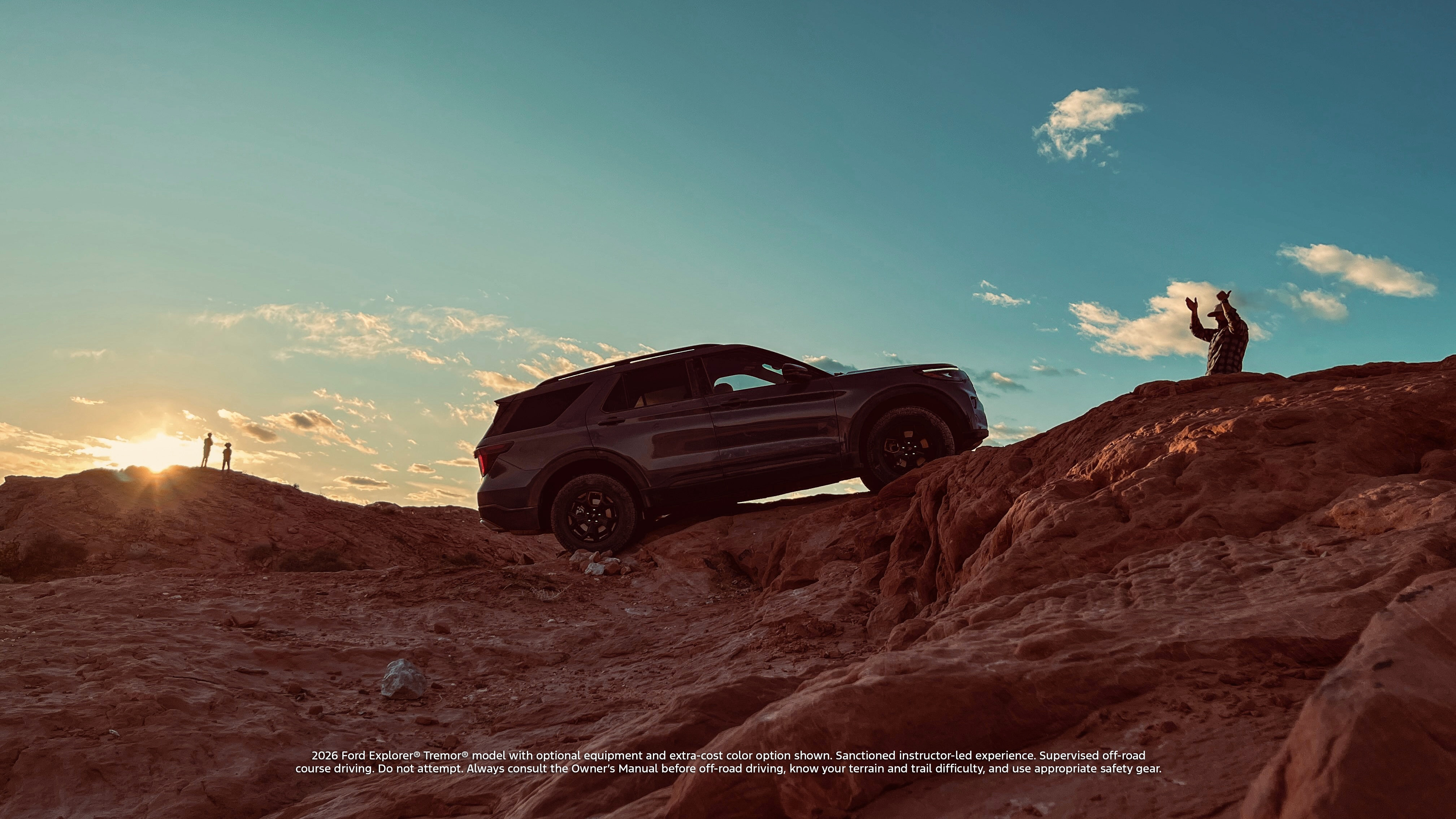 A Ford Explorer drives over an off-road trail as the sun shines over a rocky horizon. A man standing in front of the vehicle guides the driver.
